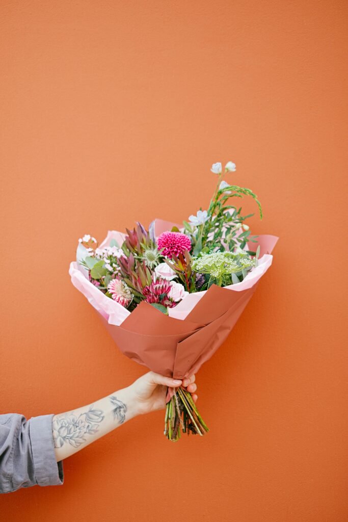 pexels-photo-5750174-5750174 Unrecognizable tattooed person with colorful various flowers with long stems wrapped in paper in hand on orange background on street